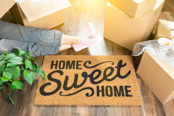 Woman Wearing Sweats Relaxing Near Home Sweet Home Welcome Mat, Moving Boxes and Plant. Woman Wearing Sweats Relaxing Near Home Sweet Home Welcome Mat, Moving Boxes and Plant. move house stock pictures, royalty-free photos & images